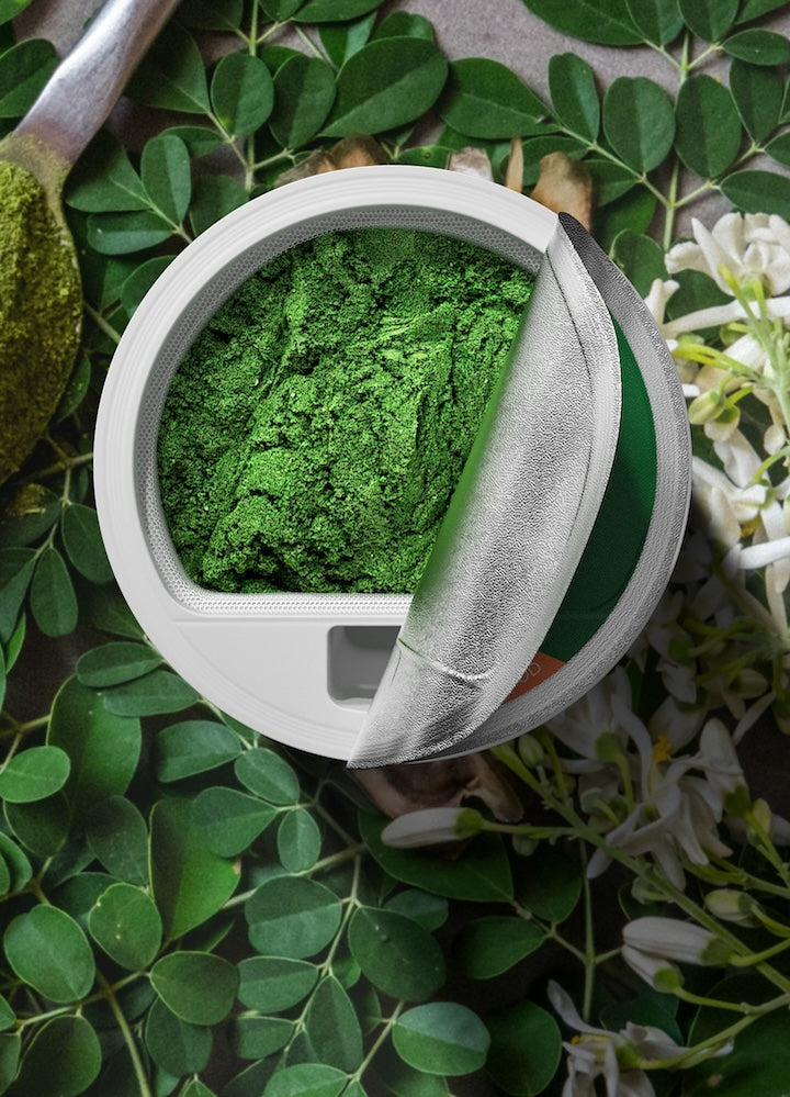 A filter flew pod  brimming with vibrant green superfood powder, set against a backdrop of fresh moringa leaves and delicate white flowers, encapsulating the natural and potent wellness boost of the FLEX Super Greens line.